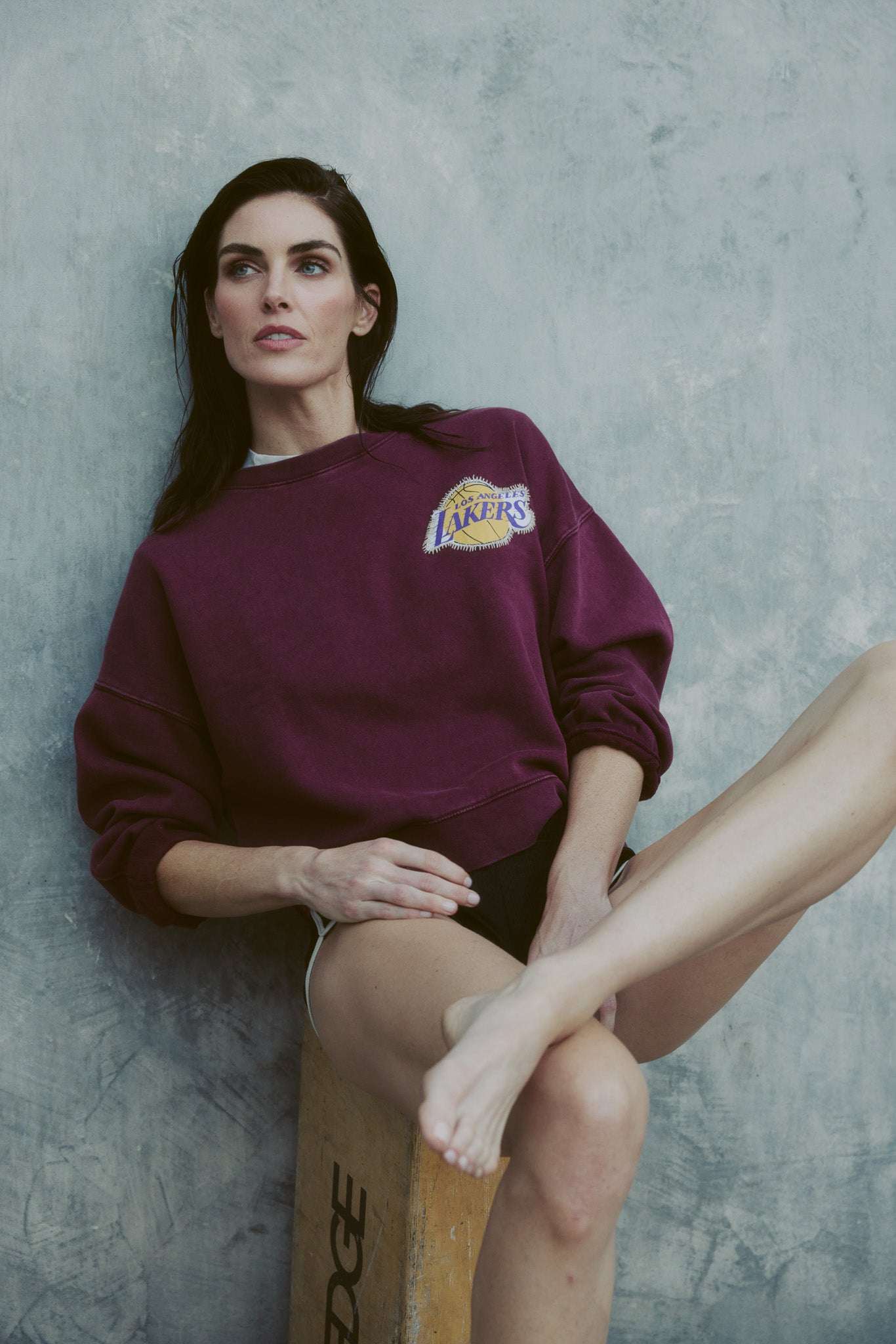 woman wearing purple Lakers Whip Stitch Crewneck sweatshirt sitting on wooden box against gray wall