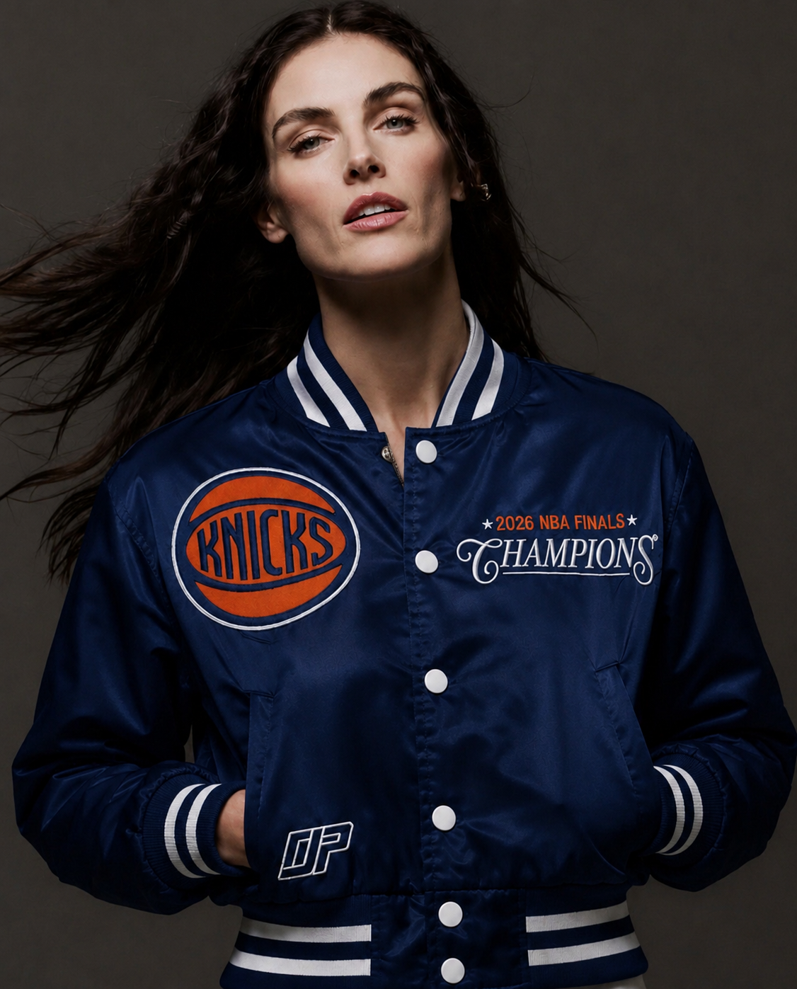 A woman wearing a navy blue satin varsity jacket with white and orange accents. The jacket features a circular 'Knicks' logo patch on the right chest and '2026 NBA Finals Champions' text on the left chest, with white buttons and striped ribbed collar, cuffs, and hem.