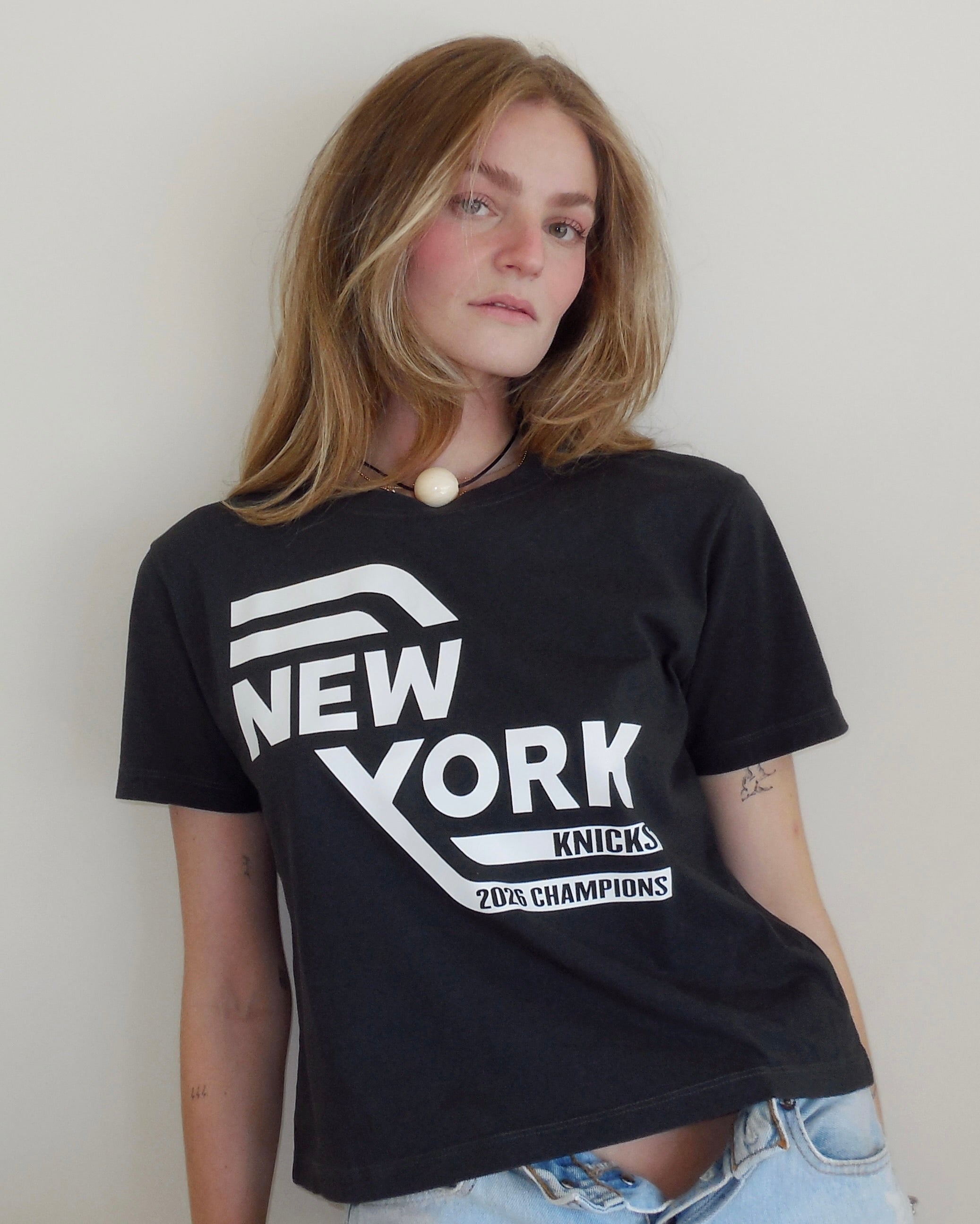 A woman with shoulder-length light brown hair wearing a black graphic t-shirt that reads 'NEW YORK KNICKS 2006 CHAMPIONS' in bold white letters, paired with light-wash blue jeans, standing against a plain off-white wall.