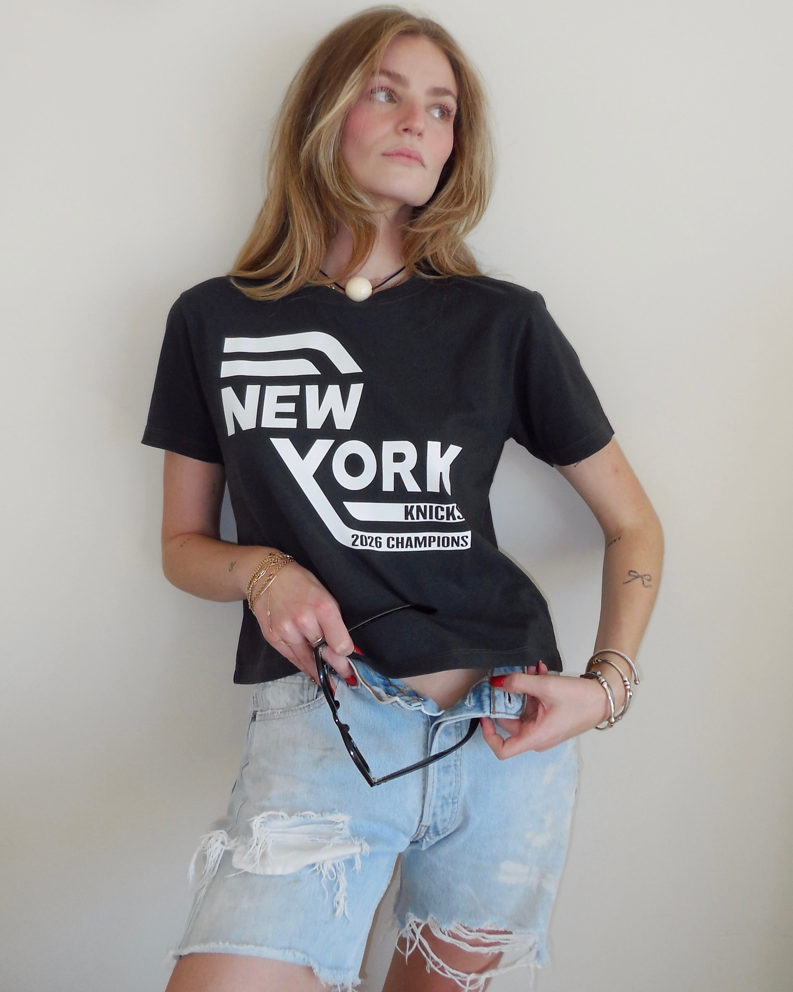 A woman wearing a black graphic t-shirt with a white 'New York Knicks' logo and light-wash distressed denim shorts, holding a pair of sunglasses against a plain off-white wall.