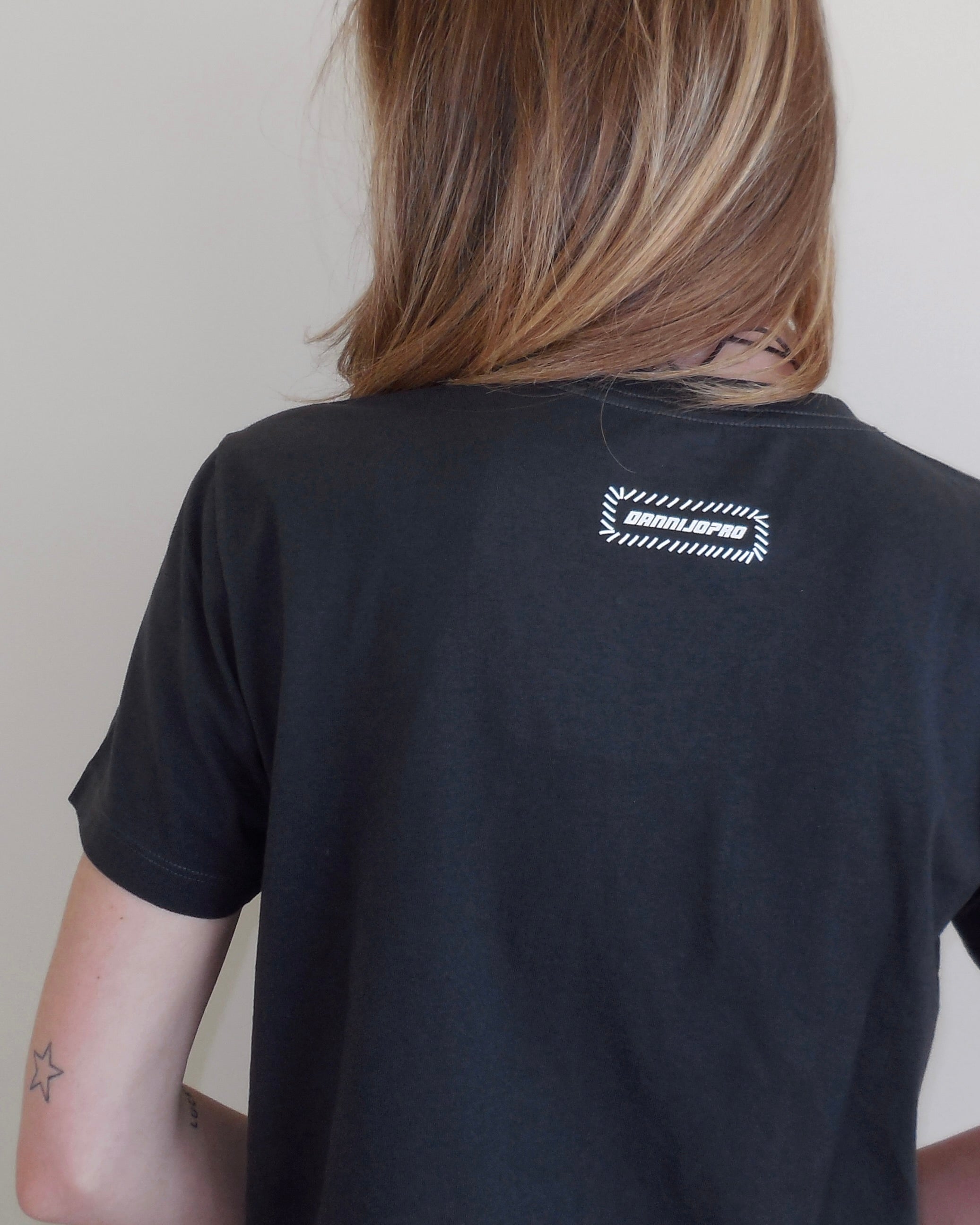 A person shown from the back wearing a black short-sleeve t-shirt with a white graphic logo on the upper back. The logo features a dashed rectangular border with the text DANNIJOPRO.