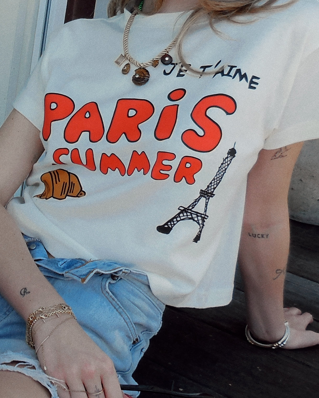 A person wearing a white graphic T-shirt with bold red 'PARIS SUMMER' text, black 'JE T'AIME' lettering, a croissant illustration, and a black Eiffel Tower graphic, paired with light blue denim shorts.