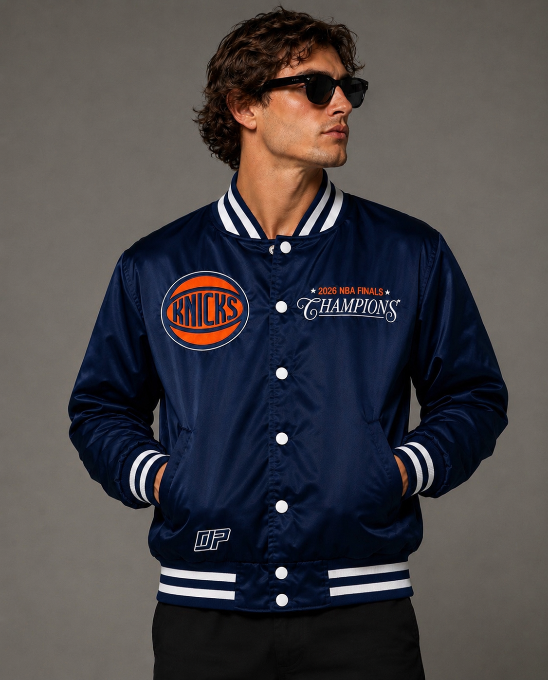 A man wearing a navy blue satin varsity jacket with white and orange details, featuring the New York Knicks logo and '2024 NBA Finals Champions' text, posed against a neutral gray background.